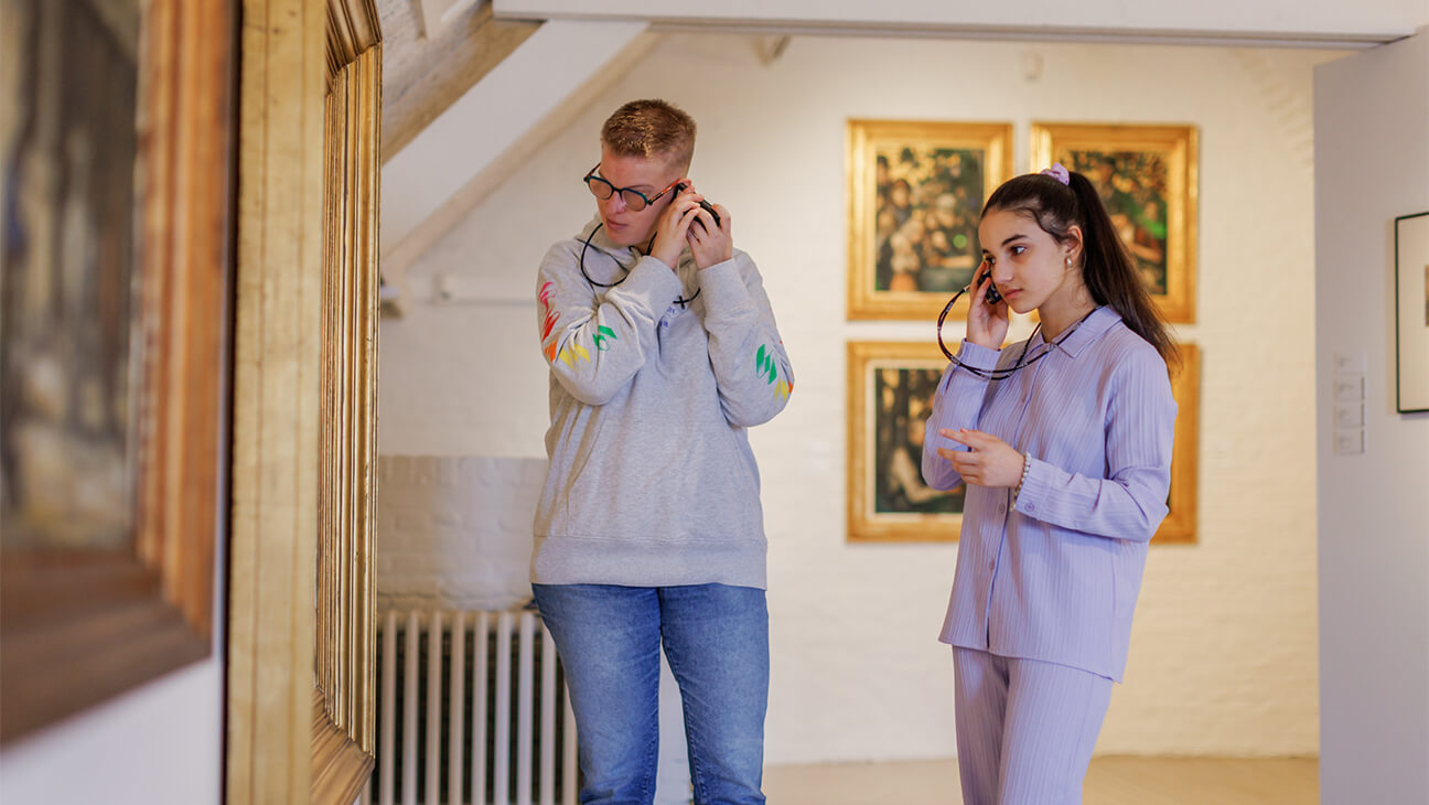 2 jongeren met een audiogids in de expo 'Bezoekers kijken naar een schilderij tijdens de expo 'Terug naar de Kempen'